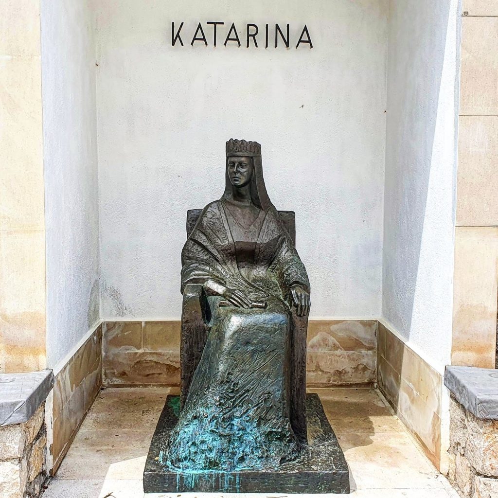 Brončani kip kraljice Katarine u sjedećem položaju uz bijeli zid